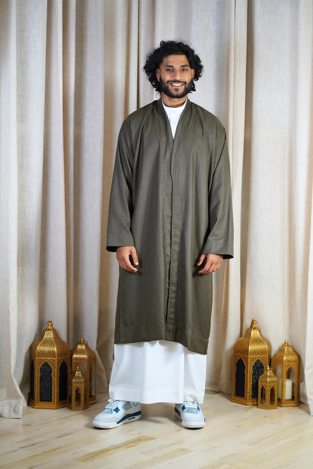 Full-length photo of a man wearing an Olive Khaki Turkish Coat layered open over a white Thobe, paired with white and blue sneakers. This is a premium modest outerwear piece in an earthy tone.