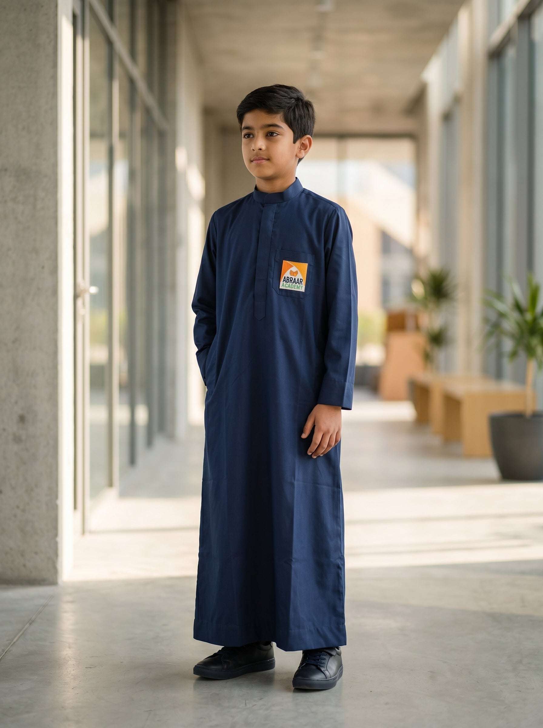 A young boy wearing the official Abraar Academy navy blue uniform thobe featuring the school logo on the chest pocket, standing in a modern, well-lit hallway.