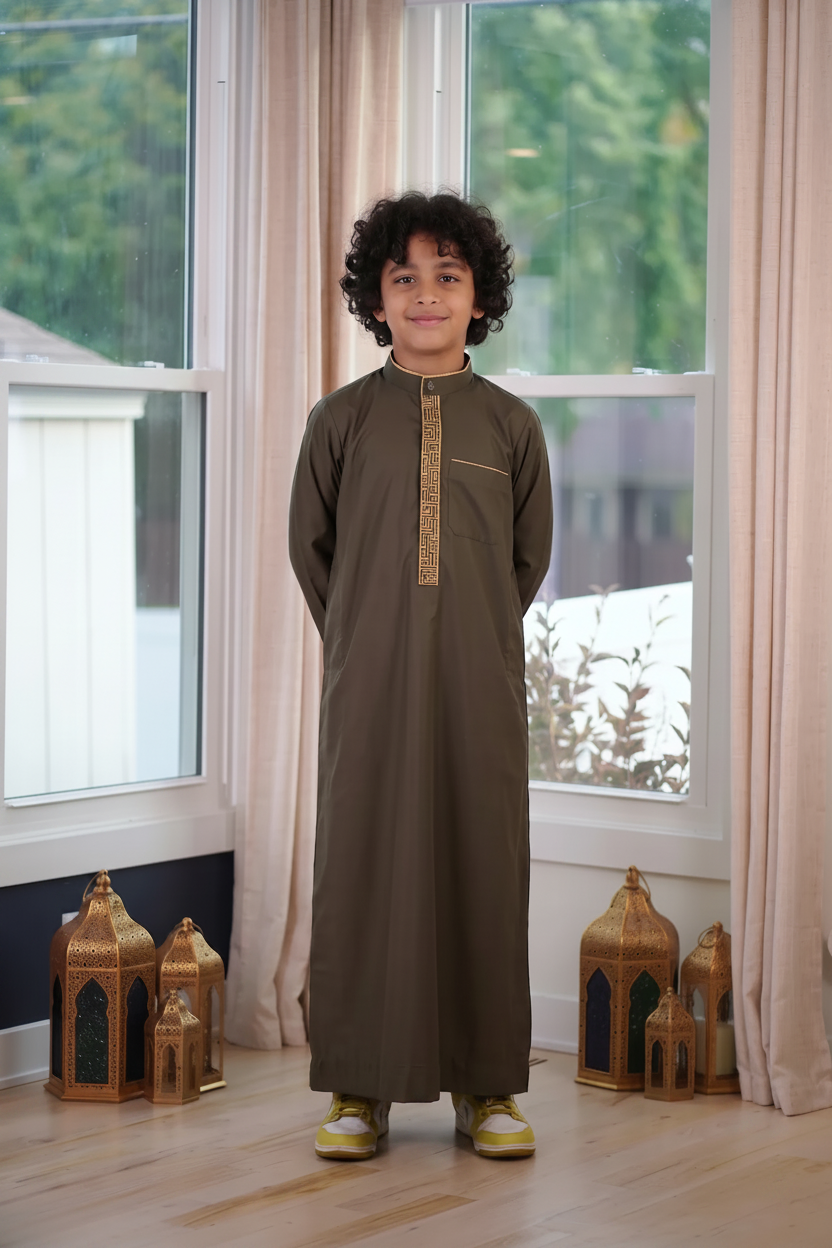 Boy wearing a premium olive green long-sleeve thobe featuring gold geometric embroidery on the placket and a mandarin collar, standing indoors next to decorative gold lanterns.