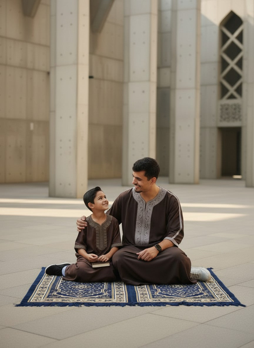 Father and son sitting together on a beautiful blue prayer mat in a modern architectural courtyard, smiling warmly at each other. Both are wearing matching premium espresso brown Moroccan thobes with elegant light grey embroidery. Spiritual Muslim family lifestyle, perfect Jumuah prayer outfits, comfortable modest menswear, and high-quality Islamic clothing for men and boys.