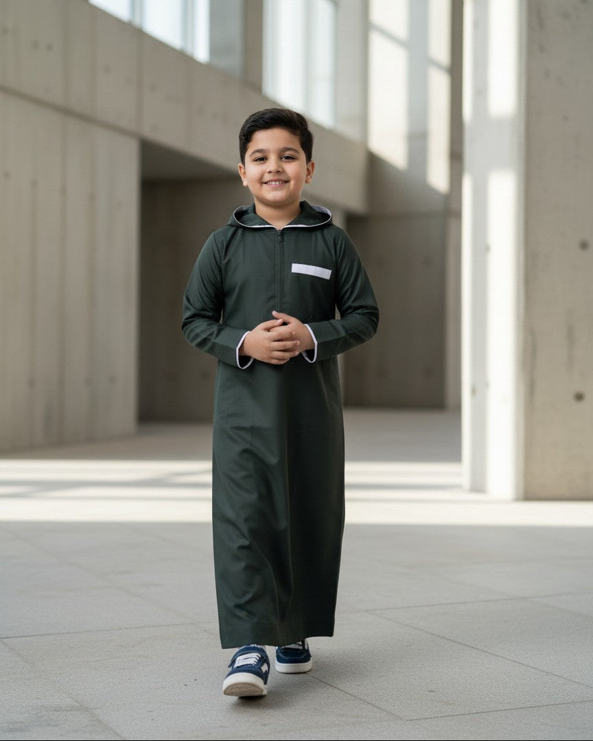 Side profile of a young boy walking through a sunny corridor wearing a dark green hooded thobe paired with casual sneakers. Versatile Islamic kids clothing, sporty boys jubba, and stylish modest streetwear.