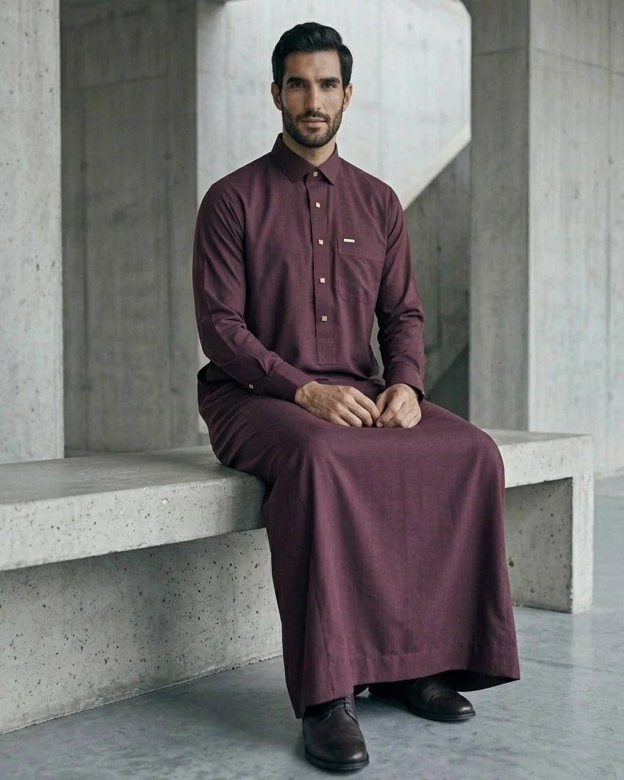 Seated pose showing full drape of premium burgundy maroon thobe with gold square buttons on concrete bench — The Topiwala wedding and Eid menswear