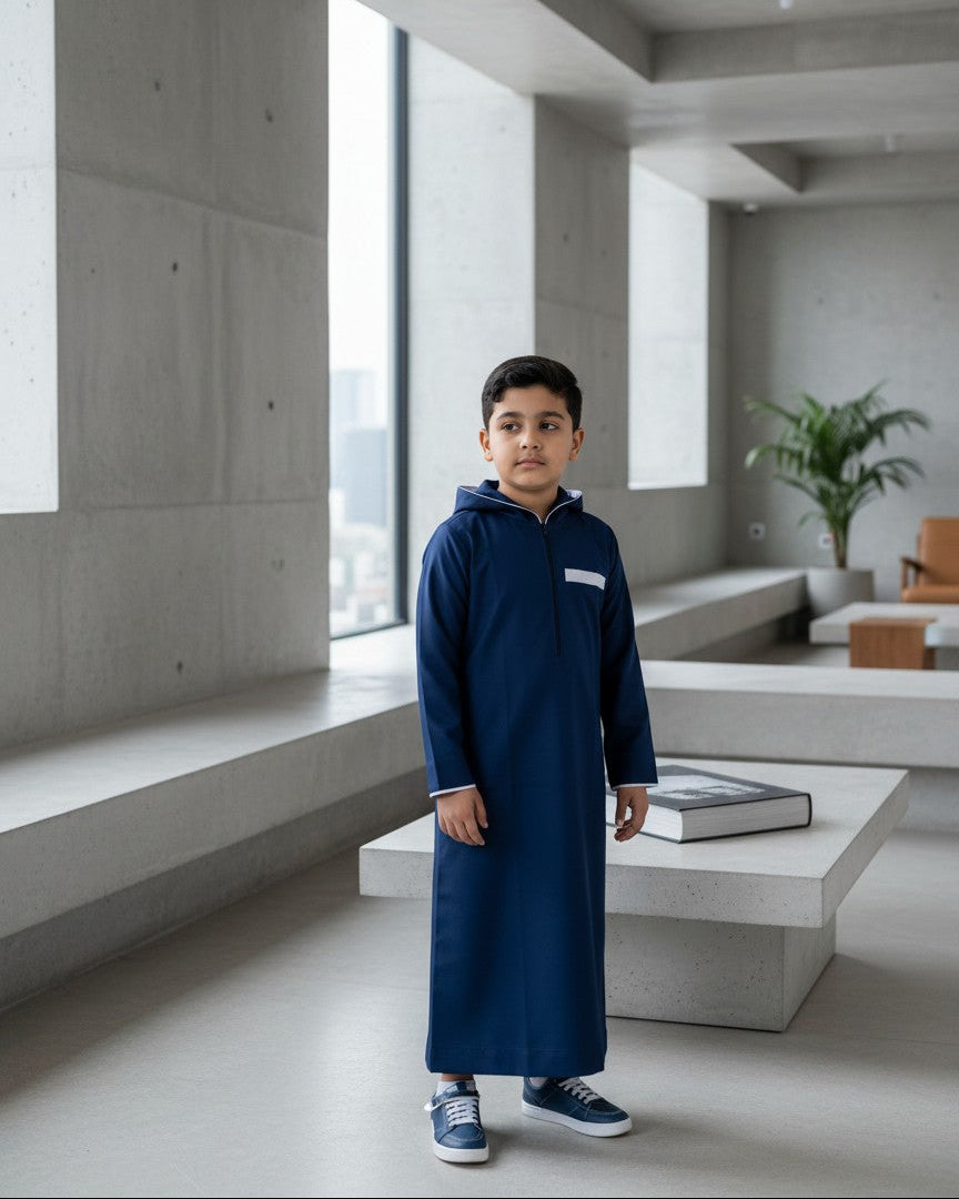 Boy standing in a modern minimalist interior wearing a sleek navy blue hooded thobe with white contrast trim. High-quality kids jubba, contemporary Muslim boys fashion, and elegant yet casual modest wear for everyday Sunnah style.