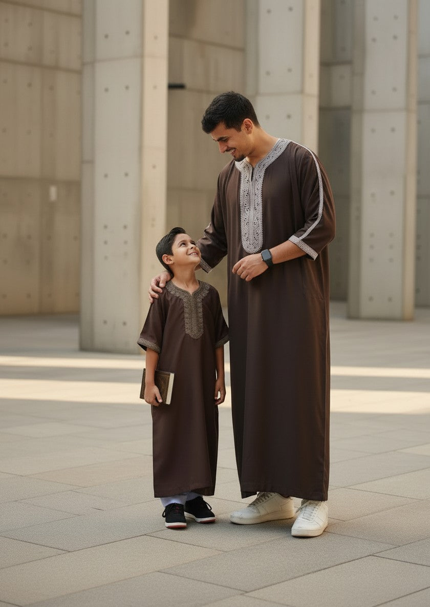 Father and son standing outdoors, smiling at each other, wearing matching premium espresso brown Moroccan thobes with light grey embroidered details. High-quality Islamic clothing, modern modest street style, beautiful family matching Eid outfits, and contemporary Sunnah fashion for men and boys.