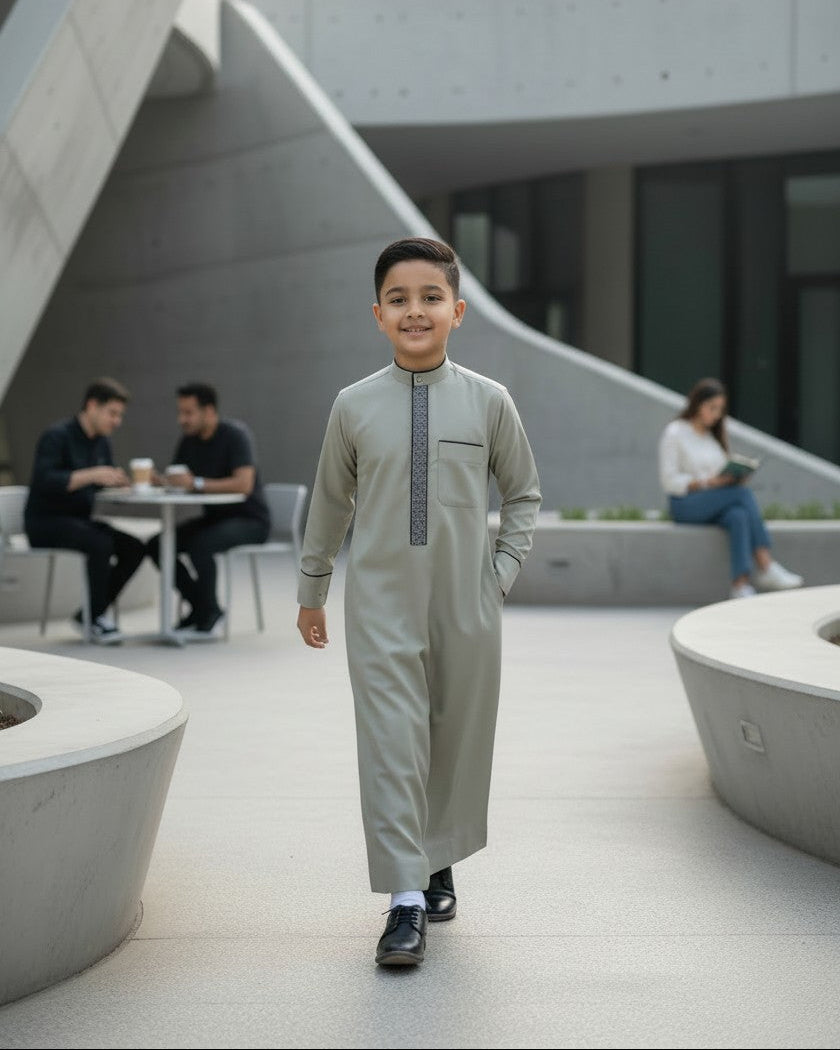 A young Muslim boy smiling and holding a tablet, wearing a premium light grey formal kids thobe with an embroidered placket. Modern Islamic children's clothing perfect for students and everyday modest wear.