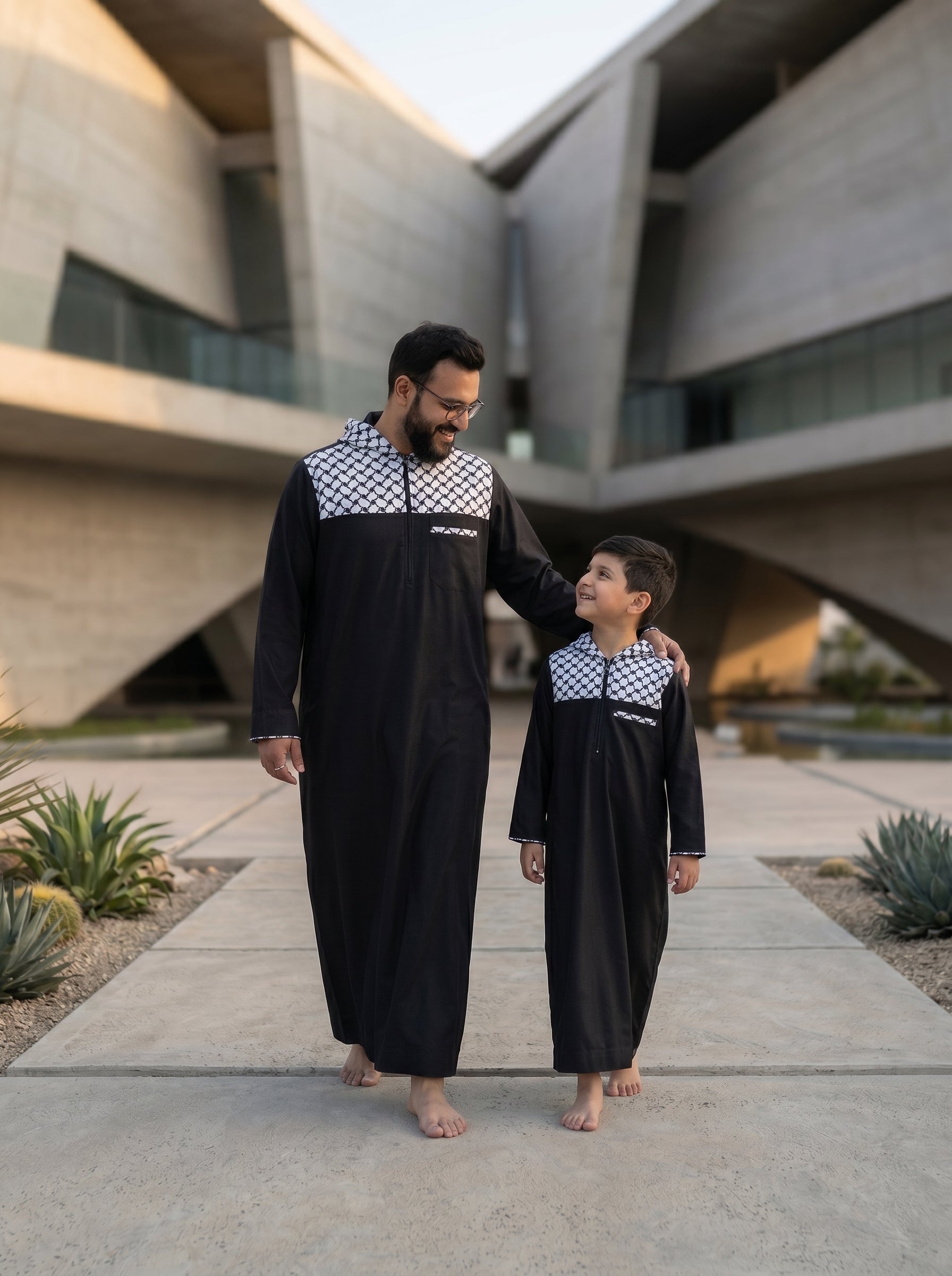 Dynamic walking shot of father and son wearing matching black hooded thobes, part of the premium collection at The Topiwala