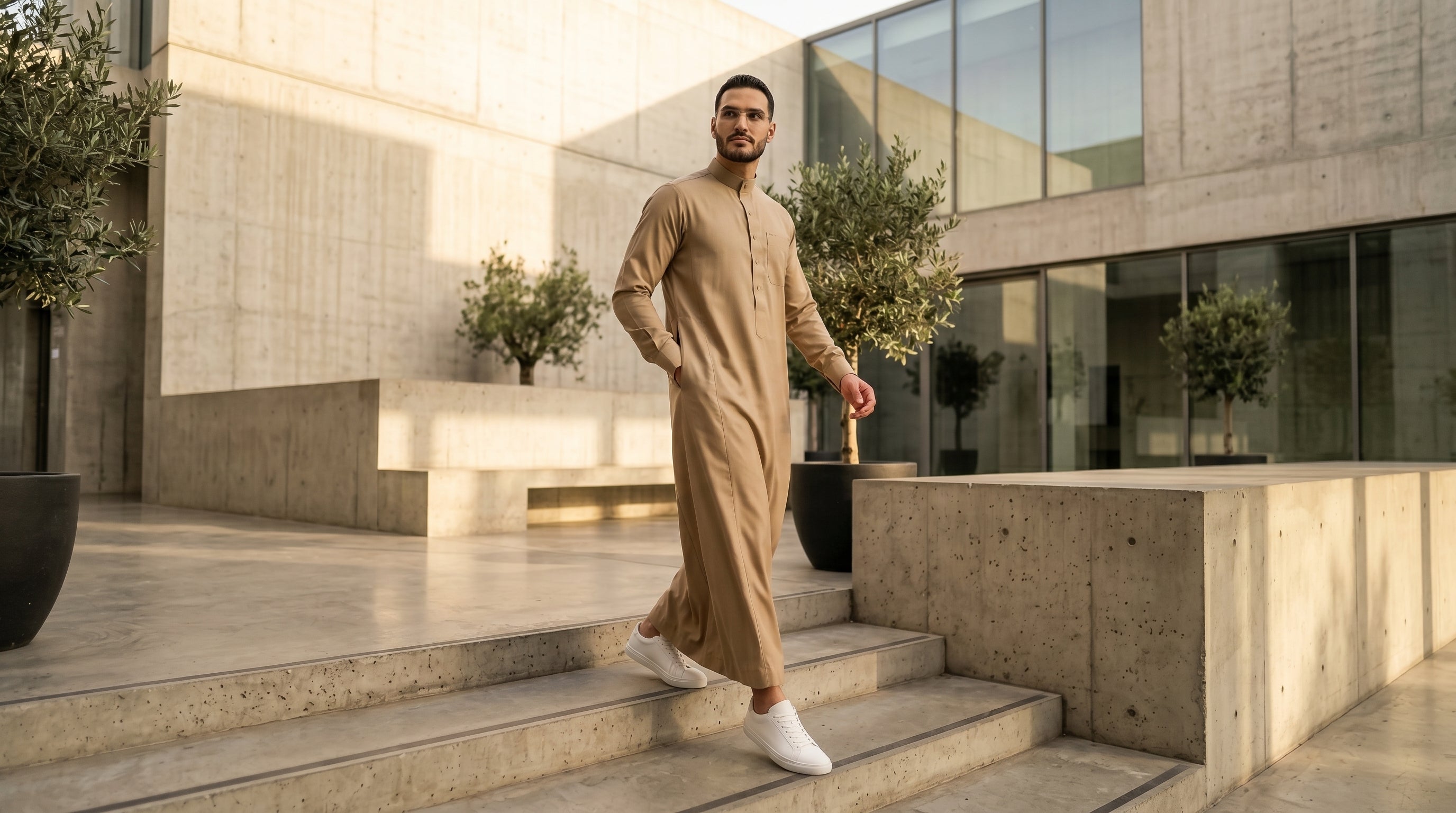 A modern Muslim man wearing a premium sand beige thobe paired with crisp white sneakers, showcasing the smart-elegant modest streetwear trend and casual everyday Sunnah style by The Topiwala.