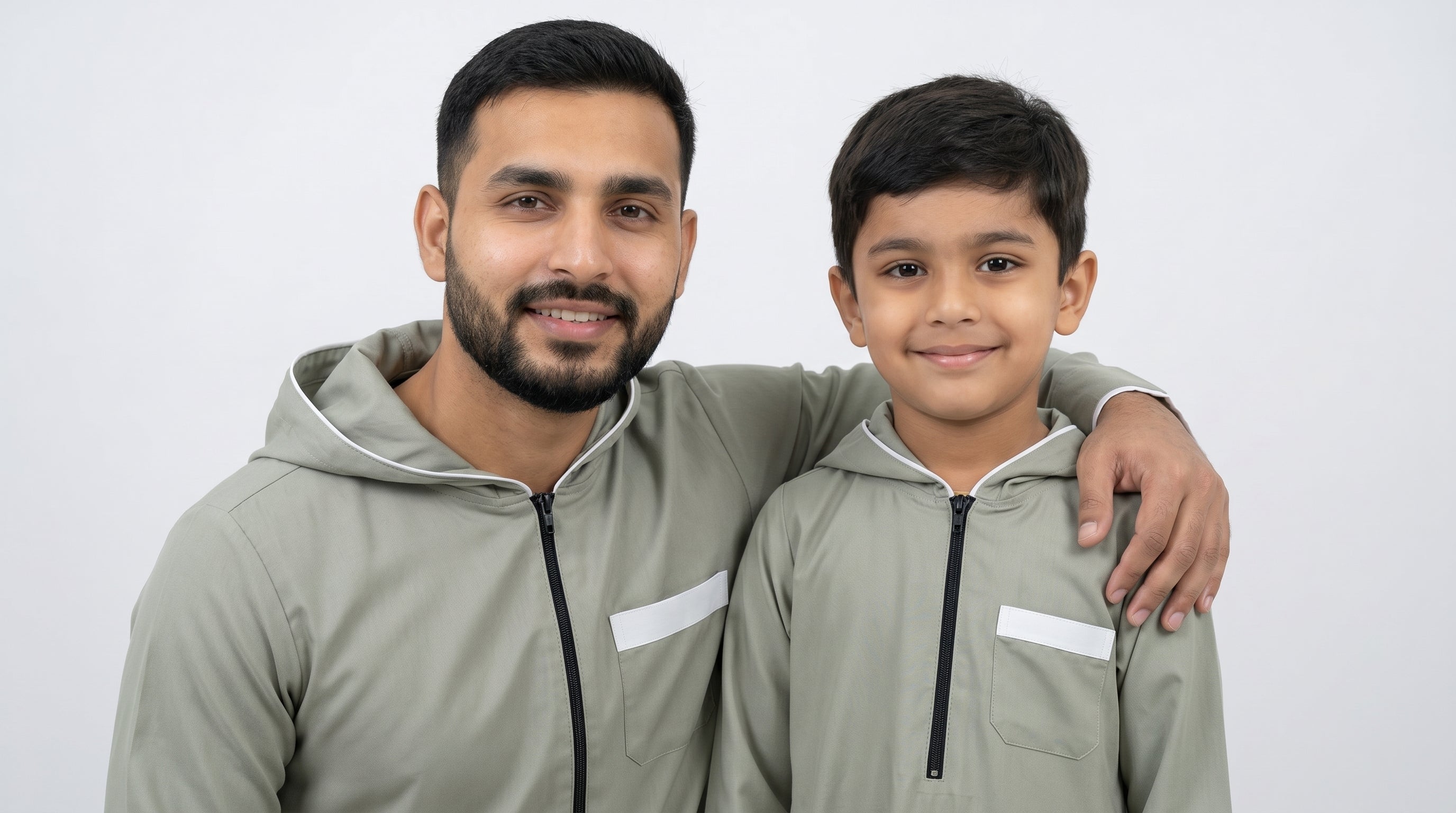 Two young Muslim boys showcasing different styles of kids jubbas: a formal sage grey embroidered thobe on the left, and a casual sporty navy blue hooded thobe on the right.