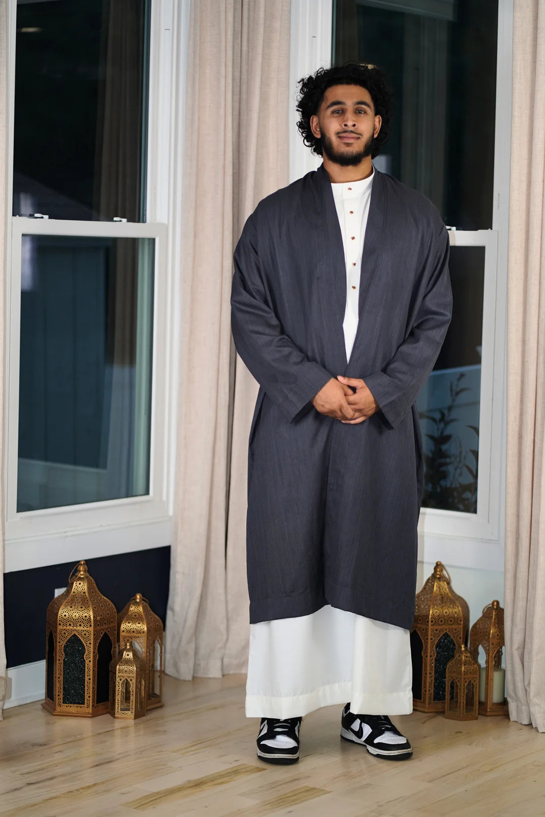 Full-length photo of a man wearing a Deep Charcoal Turkish Coat layered open over a white Thobe, paired with black and white sneakers. This is a premium modest outerwear piece for Islamic fashion.