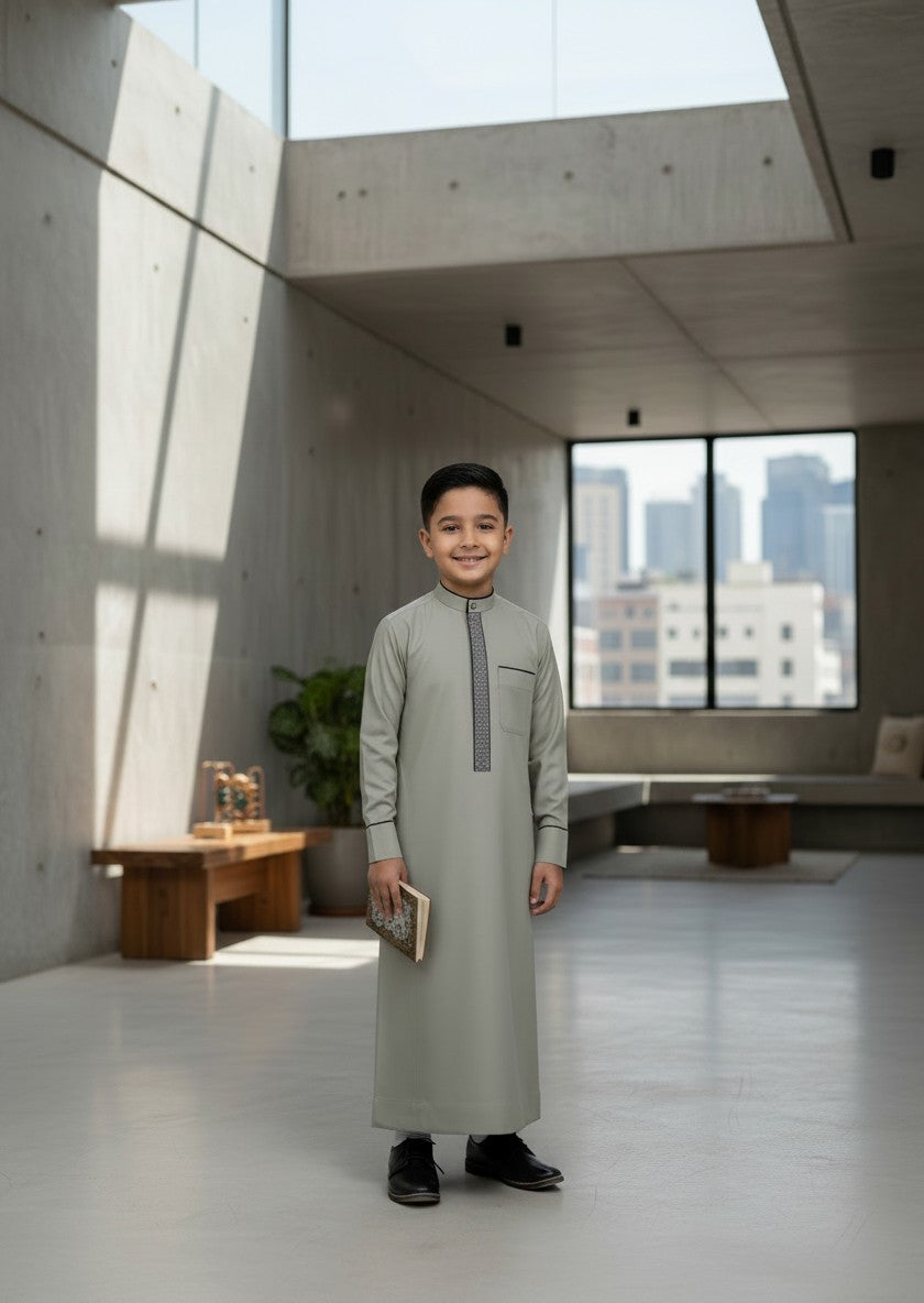 Young boy standing in a modern sunlit room holding a book, wearing an elegant light grey formal thobe with patterned embroidery. Sophisticated modest fashion for Muslim boys and premium Eid outfits.