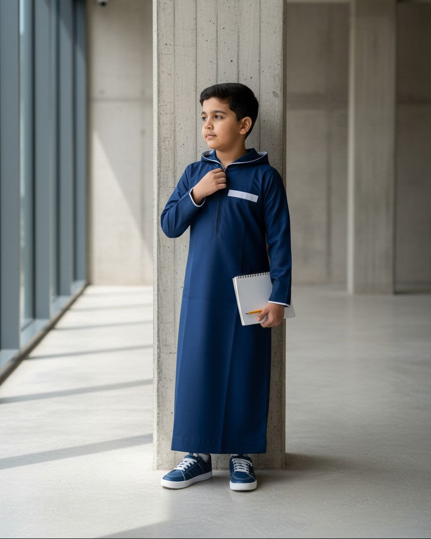 Boy sitting outdoors adjusting his navy blue hoodie-style thobe featuring white accents. Comfortable kids modest fashion, premium boys jubba, modern Islamic clothing designed for active children and everyday comfort.