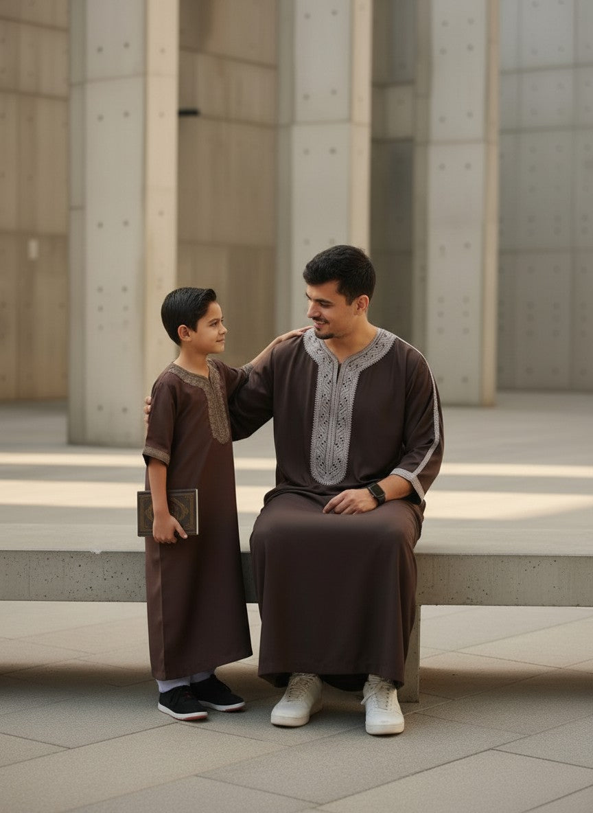 Father sitting on a concrete bench looking warmly at his son who is standing and holding a book, both wearing matching premium espresso brown Moroccan thobes with light grey embroidery. Elegant Muslim family lifestyle, comfortable kids jubba, modern modest menswear, and high-quality Islamic clothing perfect for Eid or Friday Jumuah.