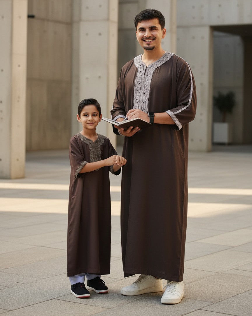 Father and son standing outdoors reading a book, both wearing matching espresso brown Moroccan-style thobes with elegant light grey embroidery. Premium Muslim menswear, comfortable kids jubba, perfect family Eid outfit, and stylish modest Islamic clothing for everyday wear.