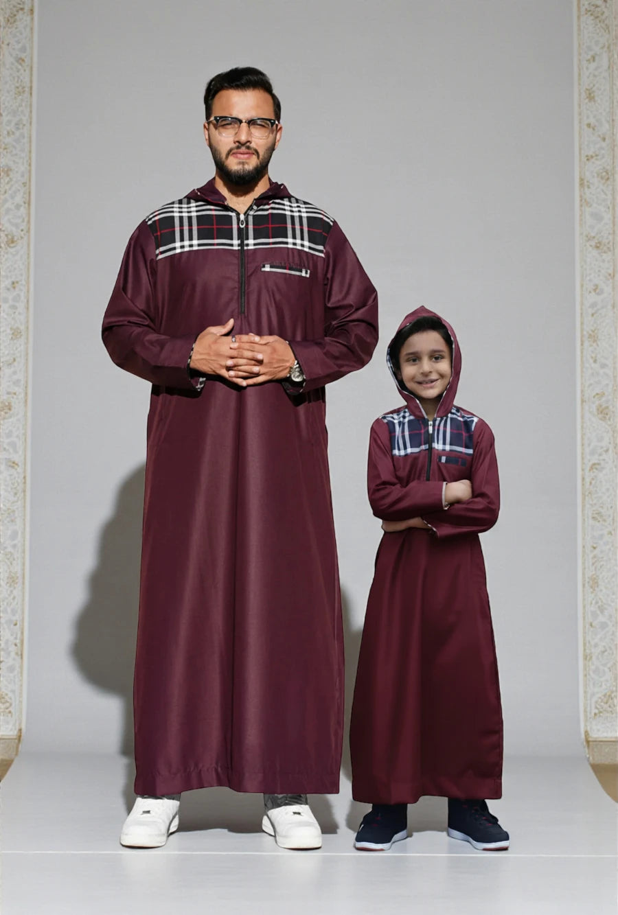 The Topi Wala matching Father and Son set featuring the Maroon Hoodie Thobe. A modern streetwear-inspired jubbah with a stylish black and white checkered (plaid) pattern covering the attached hood, shoulder yoke, and chest pocket trim. Both father and son stand side-by-side wearing sneakers, showcasing the casual, comfortable fit. Perfect for casual daily wear and urban Muslim fashion.