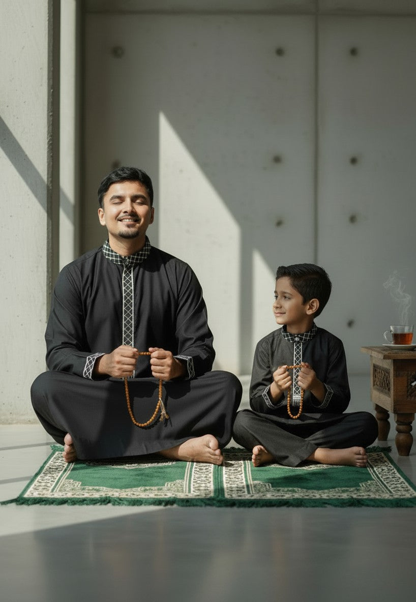 Father and son sitting comfortably on the floor in a modern minimalist interior, wearing matching premium black jubbas with black and white kufiya collar details. Relaxed Islamic clothing, elegant kids thobe, modern Muslim menswear, and stylish matching family outfits for everyday wear.