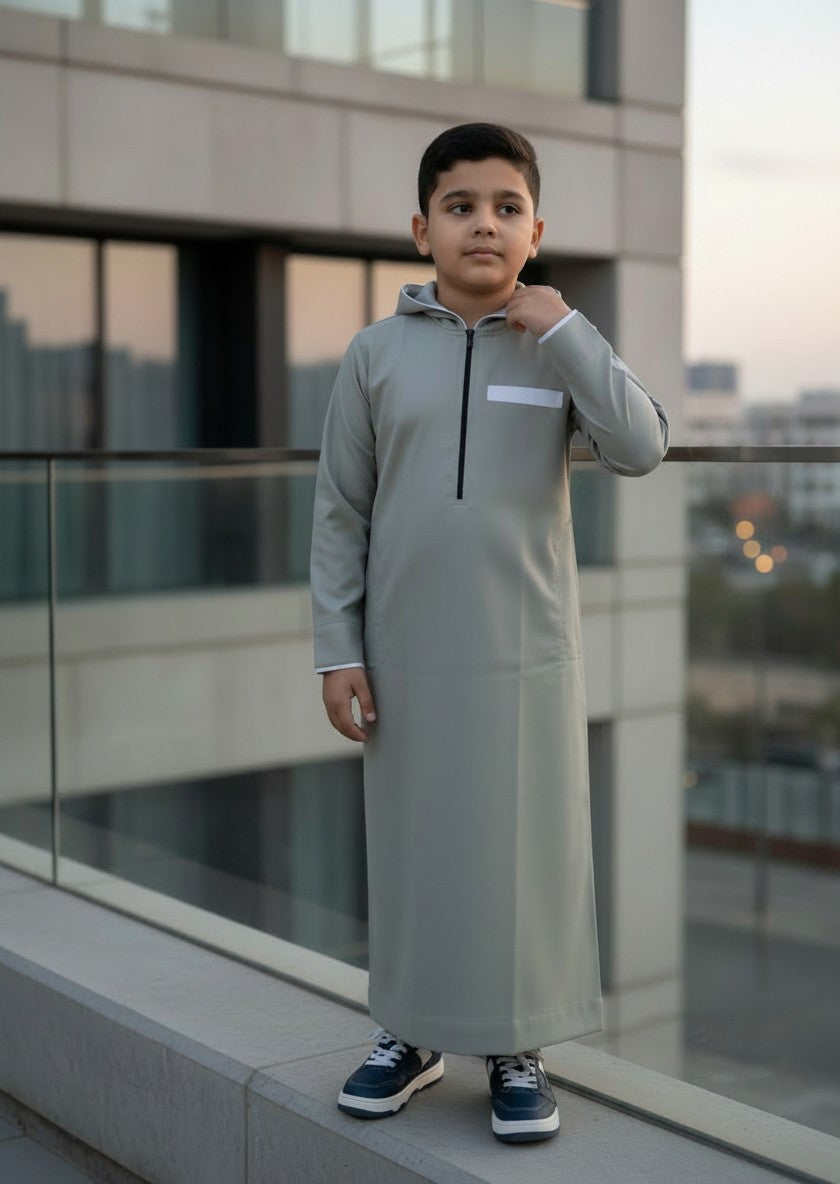 Young boy smiling and adjusting the half-zip of his premium light grey hooded thobe featuring white contrast trim. Modern sporty kids jubba and comfortable Islamic streetwear for boys.
