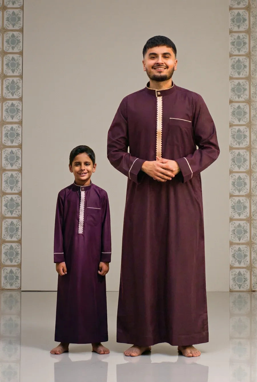 - Father and son wearing matching maroon thobes with white embroidery, standing side by side against a tiled backdrop.
