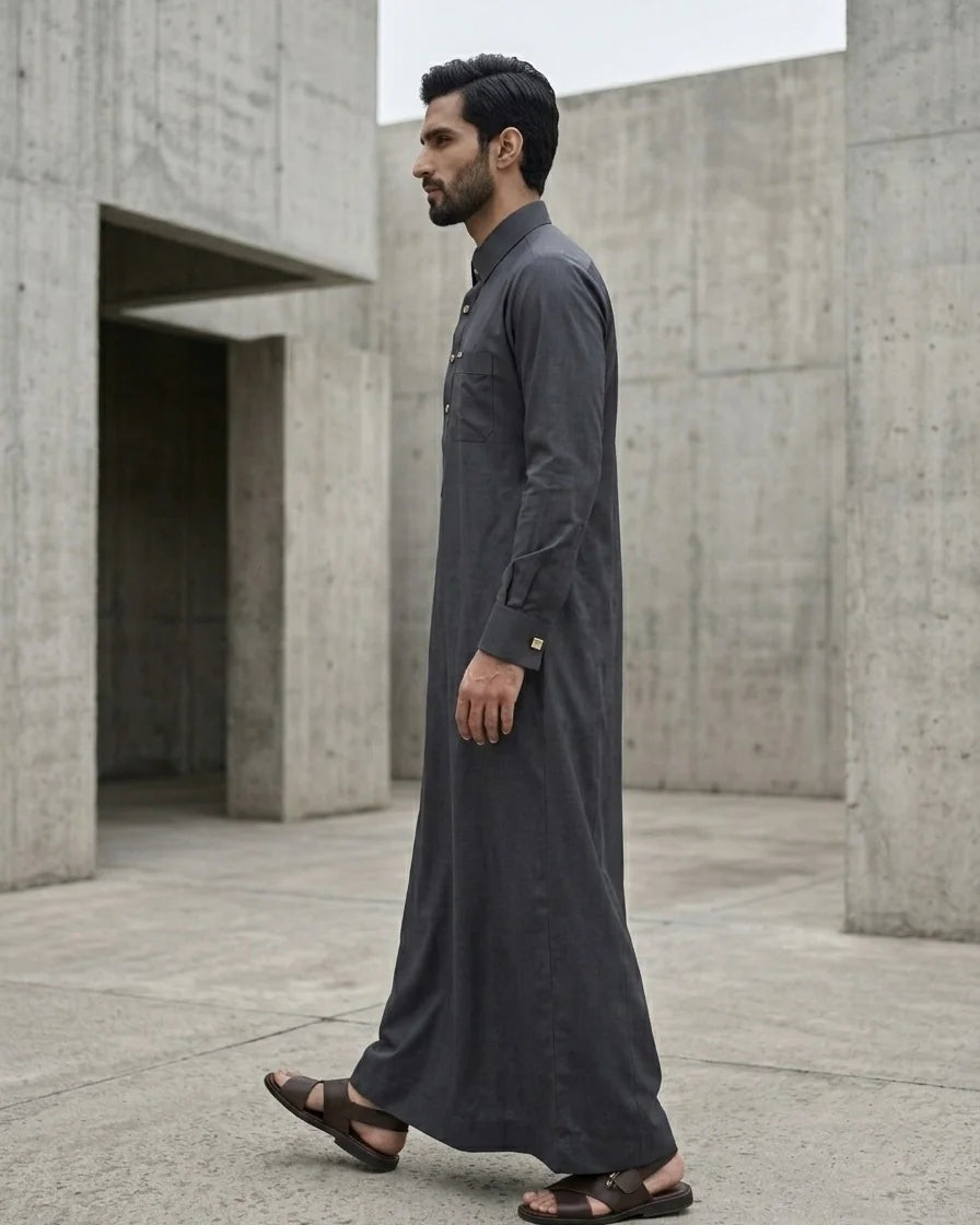 Side profile of man walking in graphite grey Saudi style thobe with structured cuffs, full length view in brutalist courtyard — The Topiwala modest menswear