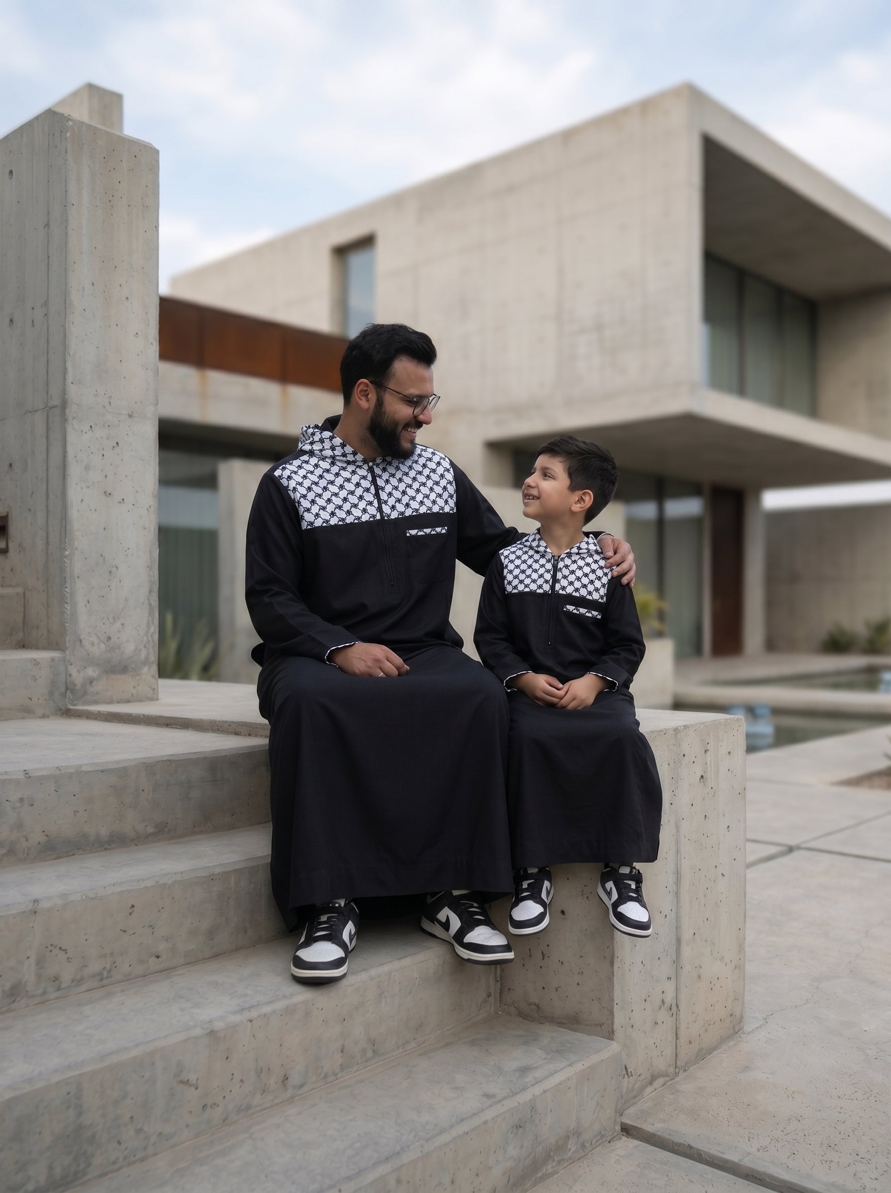 High-end editorial portrait of father and son leaning against a wall in tailored black thobes, exclusive to The Topiwala Chicago.