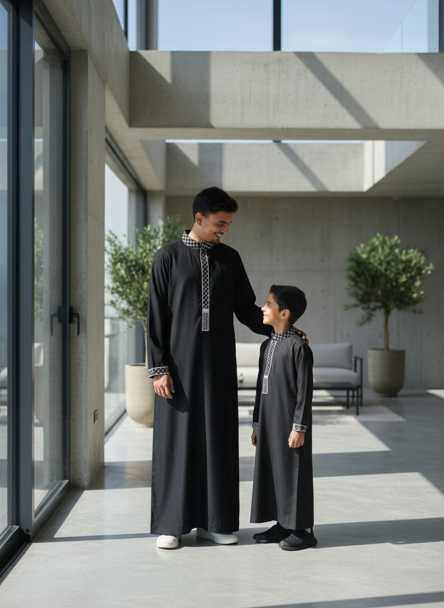 Father and son standing by a large window, smiling in matching premium black thobes with elegant black and white kufiya collar embroidery. High-quality Islamic apparel, modern modest street style, beautiful family matching Eid outfits, and contemporary Sunnah fashion for men and boys.