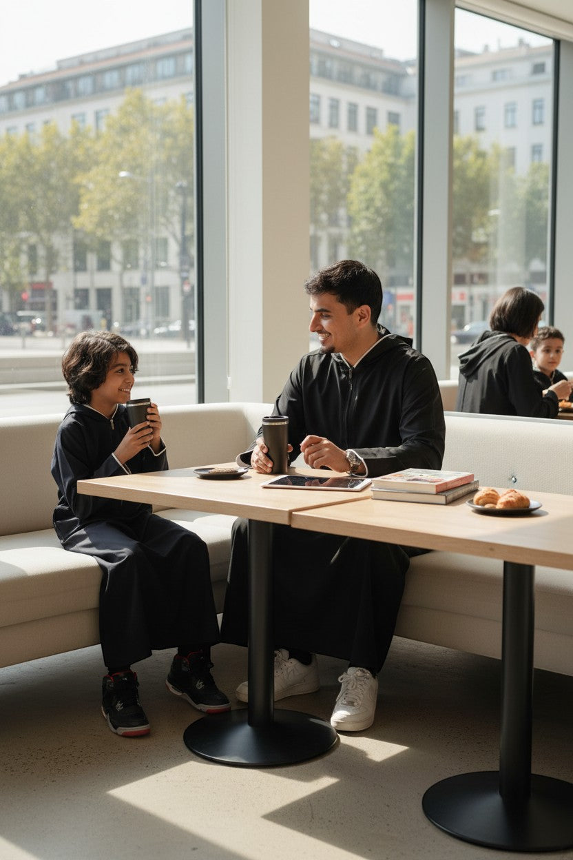 The Topi Wala father and son wearing matching black sporty hoodie thobes with white piping, sitting at a table.